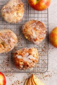 Four apple-studded pastries with glaze sit on a cooling rack, surrounded by apples, cinnamon, and apple slices on a light surface.