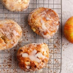 Four apple-studded pastries with glaze sit on a cooling rack, surrounded by apples, cinnamon, and apple slices on a light surface.