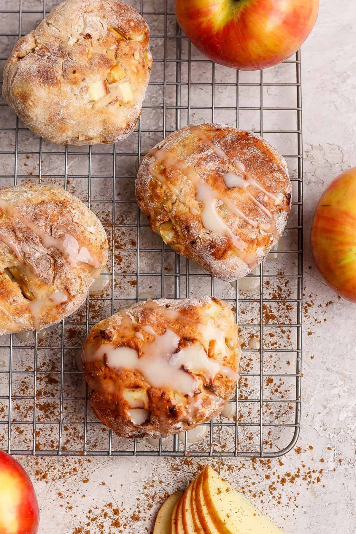 Four apple-studded pastries with glaze sit on a cooling rack, surrounded by apples, cinnamon, and apple slices on a light surface.