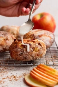 A hand drizzles white icing from a spoon onto a round baked pastry with apple pieces, resting on a cooling rack. Sliced apples and a whole apple are in the background.