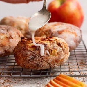 A hand drizzles white icing from a spoon onto a round baked pastry with apple pieces, resting on a cooling rack. Sliced apples and a whole apple are in the background.