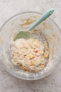 A glass bowl containing dough mixed with small apple chunks and a green spatula on a light textured surface.