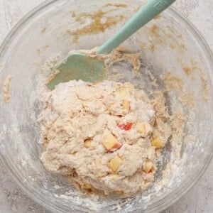 A glass bowl containing dough mixed with small apple chunks and a green spatula on a light textured surface.