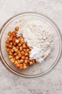 A glass bowl with diced apples coated in cinnamon on one side and a mixture of flour and white powder, likely baking powder, on the other side, on a light textured surface.