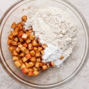 A glass bowl with diced apples coated in cinnamon on one side and a mixture of flour and white powder, likely baking powder, on the other side, on a light textured surface.