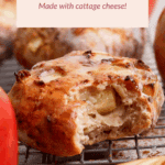 A glazed apple fritter with a bite taken out, surrounded by fresh apples and thin apple slices, on a cooling rack. Text overlay promotes high-protein apple fritters made with cottage cheese.