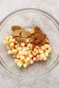 A glass bowl containing diced apples, brown sugar, and ground cinnamon on a light textured surface.
