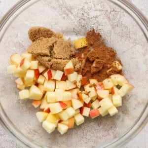 A glass bowl containing diced apples, brown sugar, and ground cinnamon on a light textured surface.