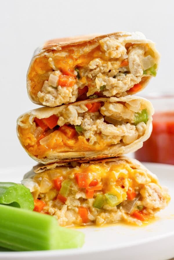 Three halves of a burrito filled with scrambled eggs, cheese, and diced vegetables are stacked on a plate next to celery sticks.