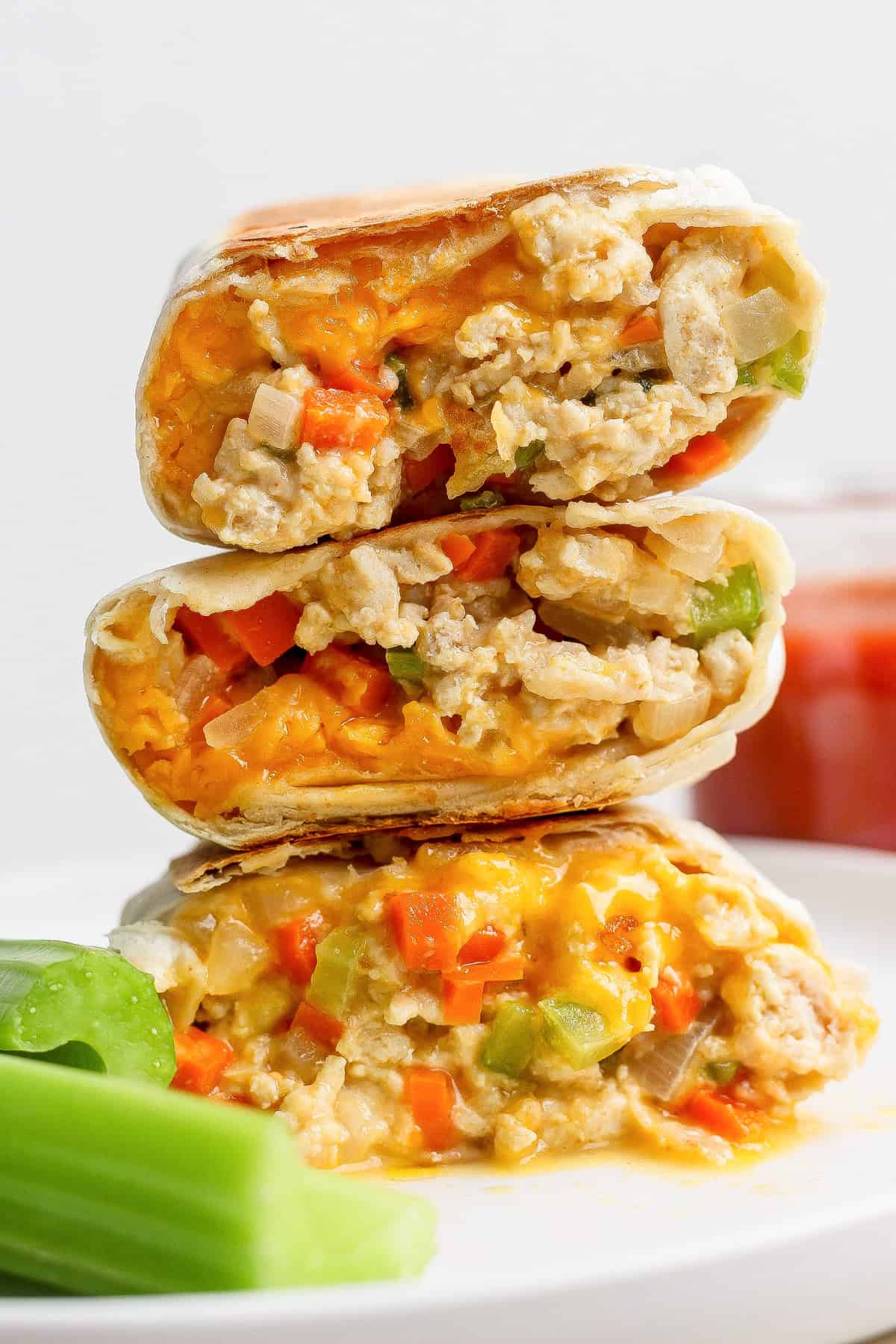Three halves of a burrito filled with scrambled eggs, cheese, and diced vegetables are stacked on a plate next to celery sticks.