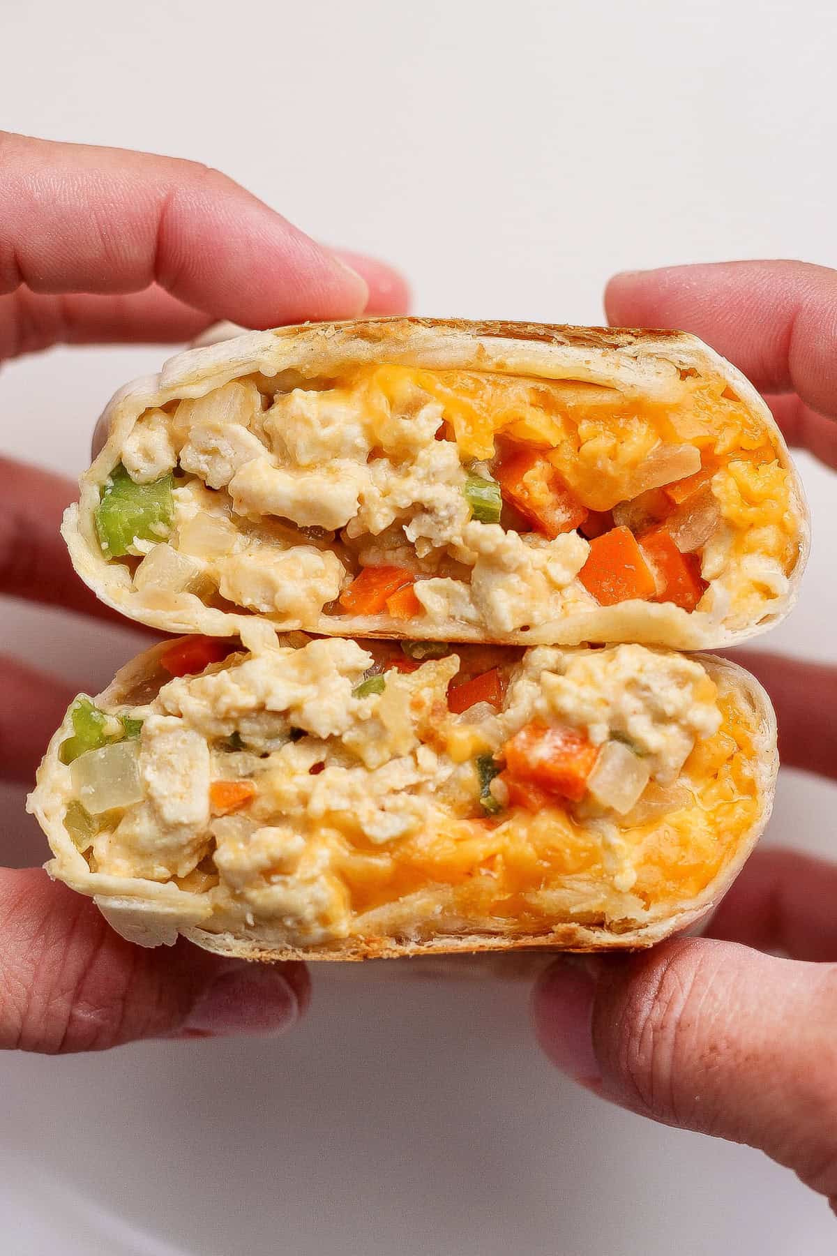 Two hands hold a breakfast burrito cut in half, showing scrambled eggs, cheddar cheese, and diced vegetables inside a tortilla wrap.
