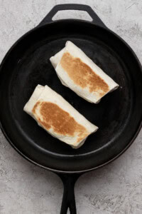 Two folded burritos being toasted in a black cast iron skillet on a light textured surface.