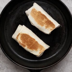Two folded burritos being toasted in a black cast iron skillet on a light textured surface.