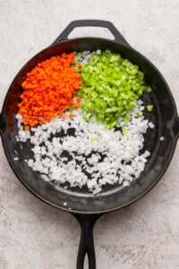 A cast iron skillet containing diced carrots, celery, and onions arranged in separate sections on a light textured surface.