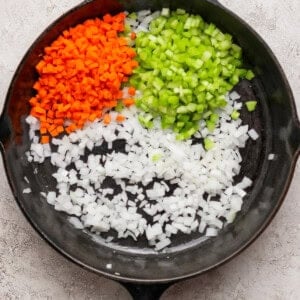 A cast iron skillet containing diced carrots, celery, and onions arranged in separate sections on a light textured surface.