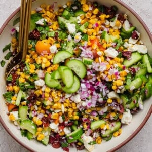 A bowl of chopped salad with corn, cucumbers, red onions, cherry tomatoes, feta cheese, and herbs, with a serving spoon.