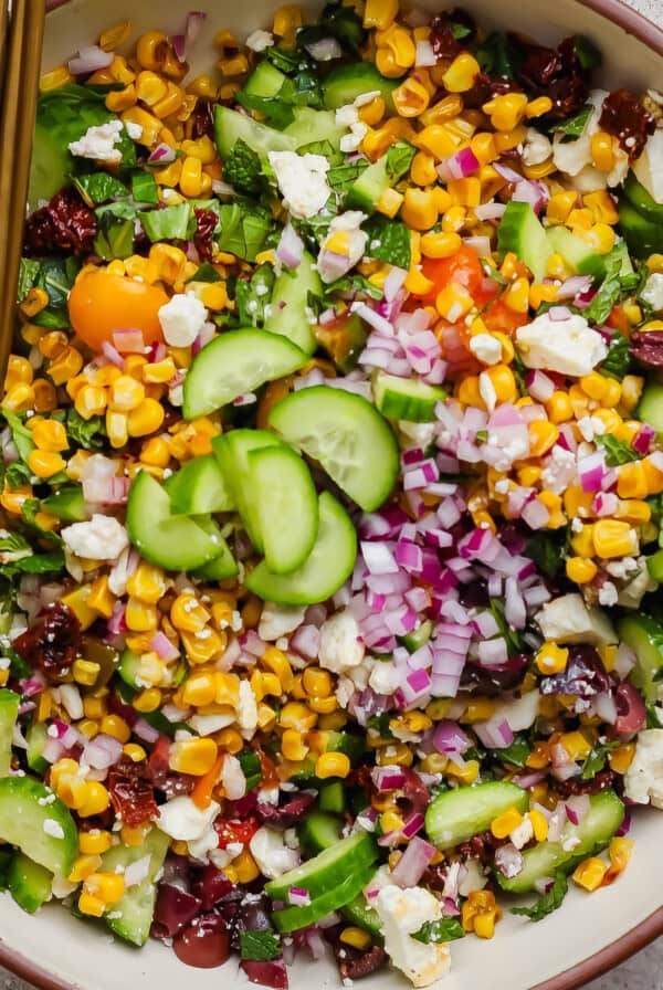 A bowl of chopped salad with corn, cucumbers, red onions, cherry tomatoes, feta cheese, and herbs, with a serving spoon.