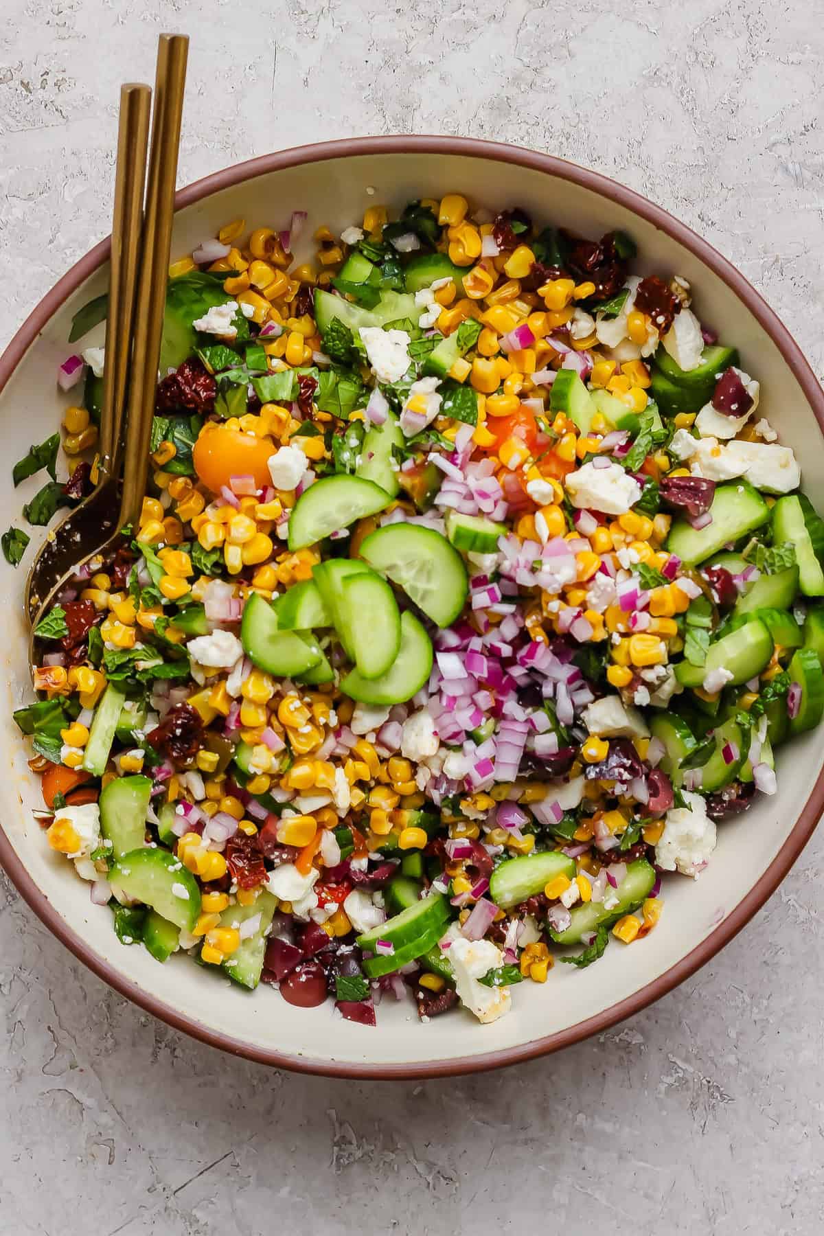 A bowl of Mediterranean corn salad with cucumbers, red onions, cherry tomatoes, feta cheese, leafy greens, and two gold forks.
