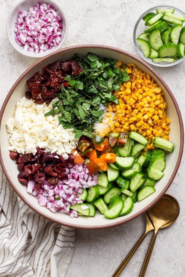 A large bowl containing chopped red onion, sun-dried tomatoes, feta cheese, olives, basil, mint, corn, cherry tomatoes, and cucumber, with bowls of onions and cucumbers on the side.
