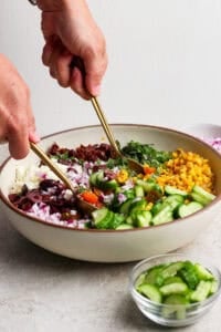 A person prepares a mediterranean corn salad, mixing chopped cucumber, corn, red onion, olives, sun-dried tomatoes, greens, and feta cheese in a large bowl using two gold utensils.