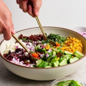 A person prepares a mediterranean corn salad, mixing chopped cucumber, corn, red onion, olives, sun-dried tomatoes, greens, and feta cheese in a large bowl using two gold utensils.