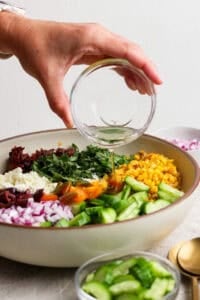 A hand pours dressing from a glass bowl onto a vibrant Mediterranean corn salad featuring cucumber, red onion, tomatoes, olives, feta cheese, and greens in a large white bowl.