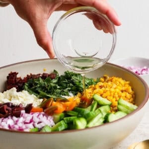 A hand pours dressing from a glass bowl onto a vibrant Mediterranean corn salad featuring cucumber, red onion, tomatoes, olives, feta cheese, and greens in a large white bowl.