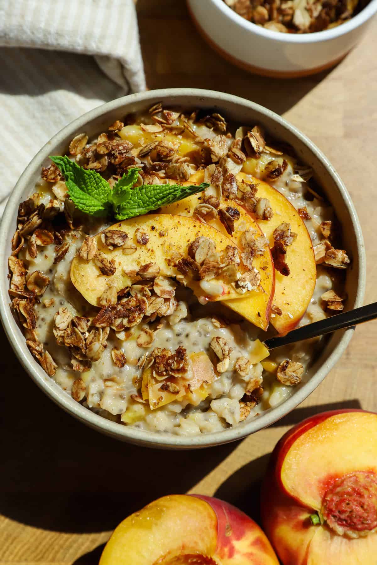 A bowl of oatmeal topped with granola, fresh peach slices, and a mint sprig, with a spoon inside and peach halves nearby on a wooden surface.