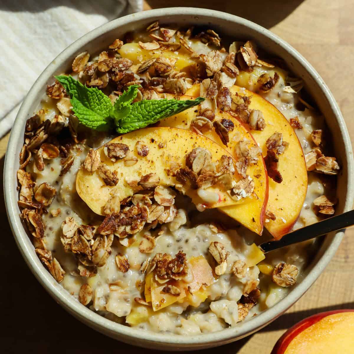 A bowl of oatmeal topped with granola, peach slices, and a sprig of mint, with a spoon placed inside.