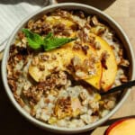 A bowl of oatmeal topped with granola, peach slices, and a sprig of mint, with a spoon placed inside.