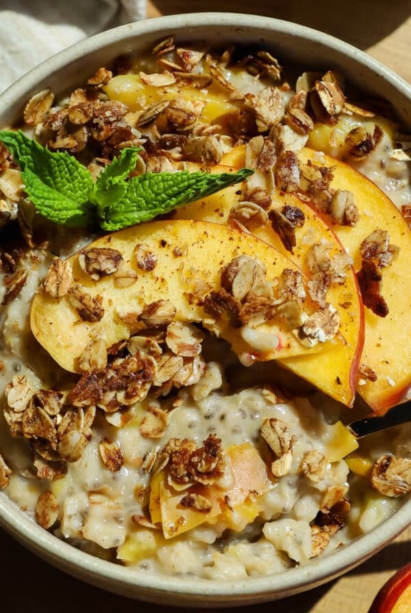 A bowl of oatmeal topped with granola, peach slices, and a sprig of mint, with a spoon placed inside.