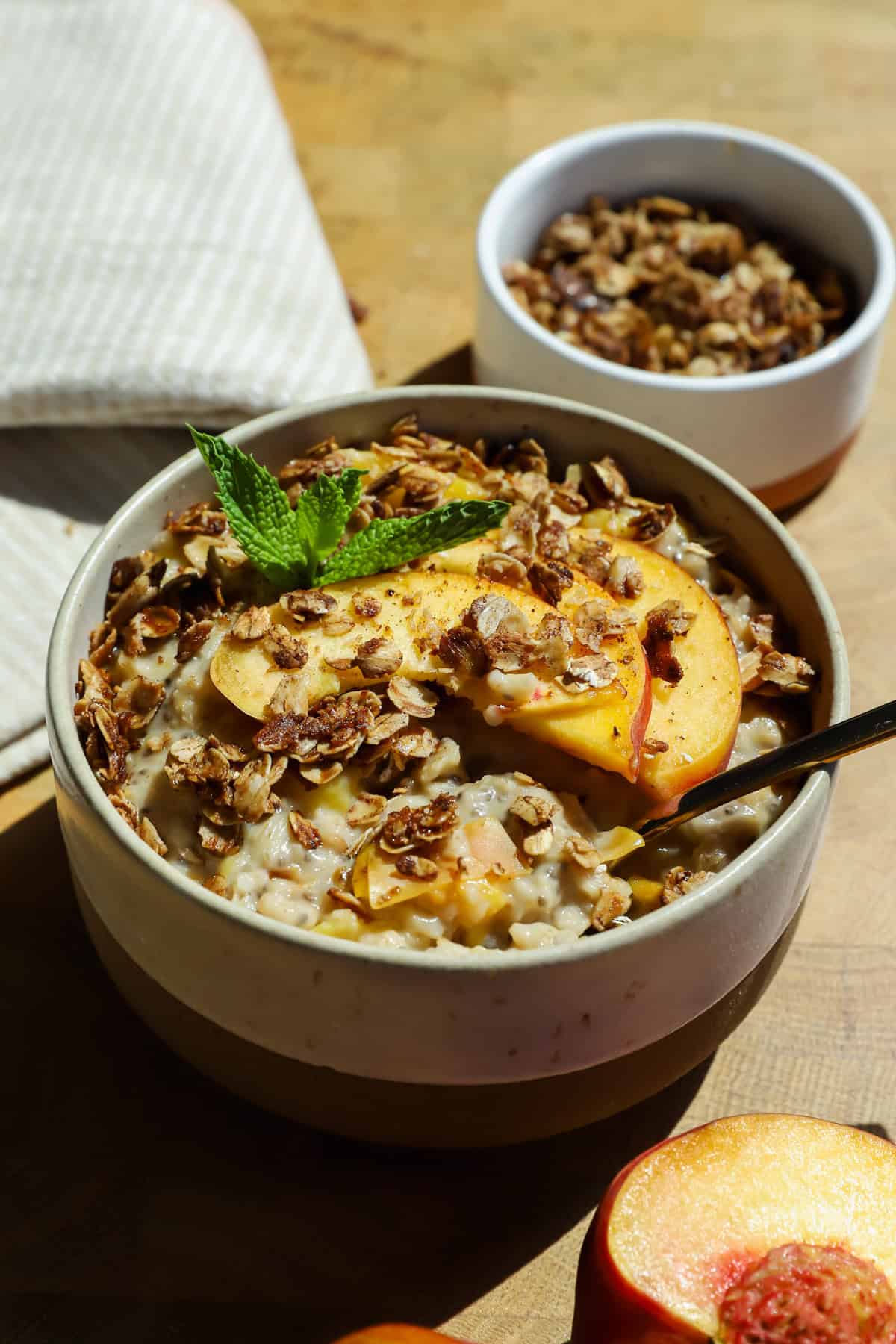 A bowl of oatmeal topped with peach slices, granola, and a mint sprig, with a spoon inside. A small bowl of granola and a peach half are nearby on a wooden surface.