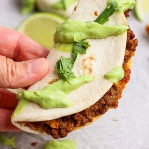 A hand holds a taco filled with ground beef and topped with green sauce and chopped cilantro on a light surface.