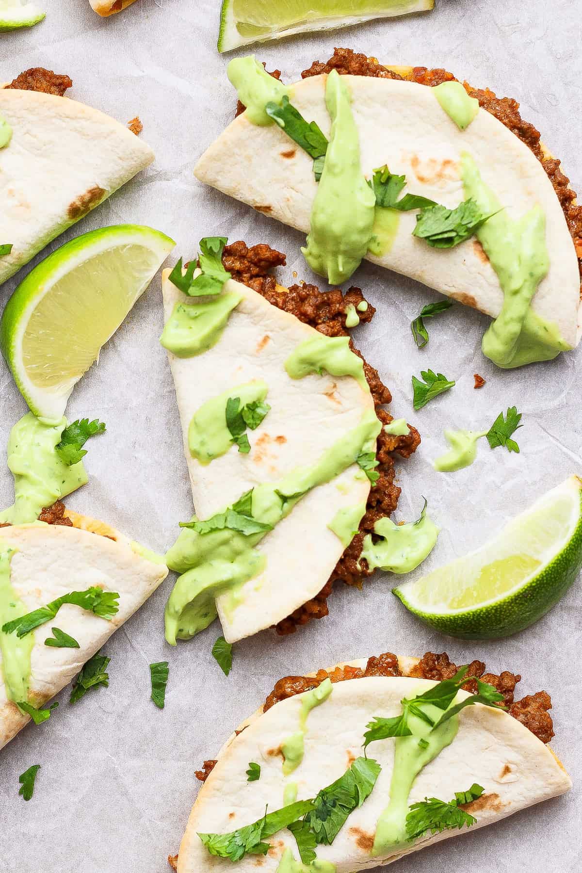 Three folded tacos filled with ground beef, topped with green sauce and fresh cilantro, are arranged on parchment paper with lime wedges.