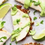 Taco halves with ground meat filling, topped with green sauce and cilantro, served with lime wedges on a white surface.