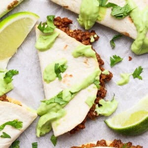 Taco halves with ground meat filling, topped with green sauce and cilantro, served with lime wedges on a white surface.