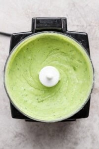 Top view of a food processor containing smooth, creamy green sauce or dip, likely a blended avocado or herb mixture.
