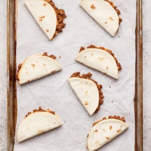 Six folded flour tortillas filled with ground beef are arranged on a parchment-lined baking sheet.