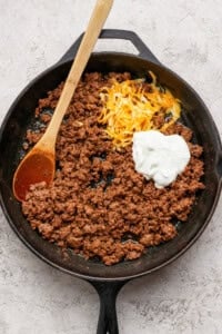 Ground beef in a cast iron skillet with shredded cheese and a dollop of sour cream, being stirred with a wooden spoon.