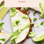 Overhead view of beef tacos on tortillas, topped with sliced avocado and chopped cilantro, with lime wedges on a parchment-lined surface.