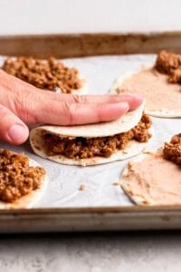 A hand folds a tortilla filled with ground meat mixture on a baking sheet lined with parchment paper, with more tortillas being assembled nearby.