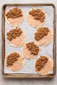 Seven tortillas spread with refried beans and ground beef mixture are arranged on a parchment-lined baking sheet.