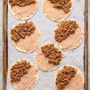 Seven tortillas spread with refried beans and ground beef mixture are arranged on a parchment-lined baking sheet.