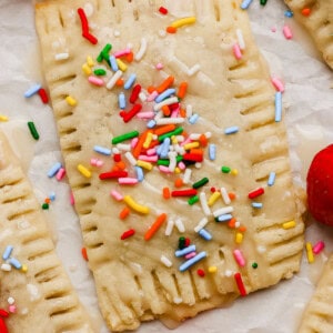 Rectangular pastry with icing and colorful sprinkles, surrounded by fresh strawberries on a white surface.