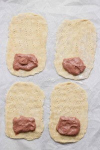 Four rectangular dough pieces, each with fork marks and a portion of meat paste on one end, are arranged on a parchment-lined surface.