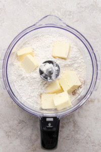 Butter chunks and flour in a food processor bowl, ready to be mixed, viewed from above on a light countertop.