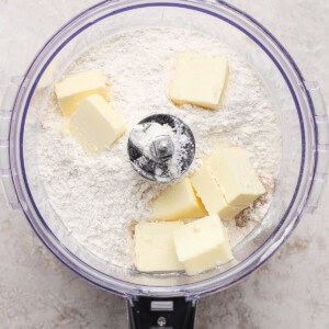Butter chunks and flour in a food processor bowl, ready to be mixed, viewed from above on a light countertop.