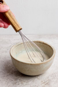 A hand holding a whisk stirring a light-colored mixture in a ceramic bowl on a light surface.