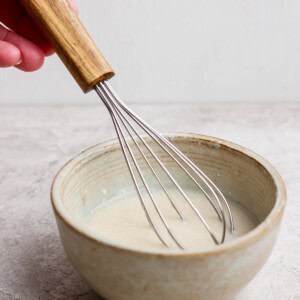 A hand holding a whisk stirring a light-colored mixture in a ceramic bowl on a light surface.
