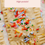 A close-up of a homemade strawberry Pop Tart with icing and colorful sprinkles, surrounded by fresh strawberries. Text on image reads: “Strawberry Pop Tarts - High-protein!”.